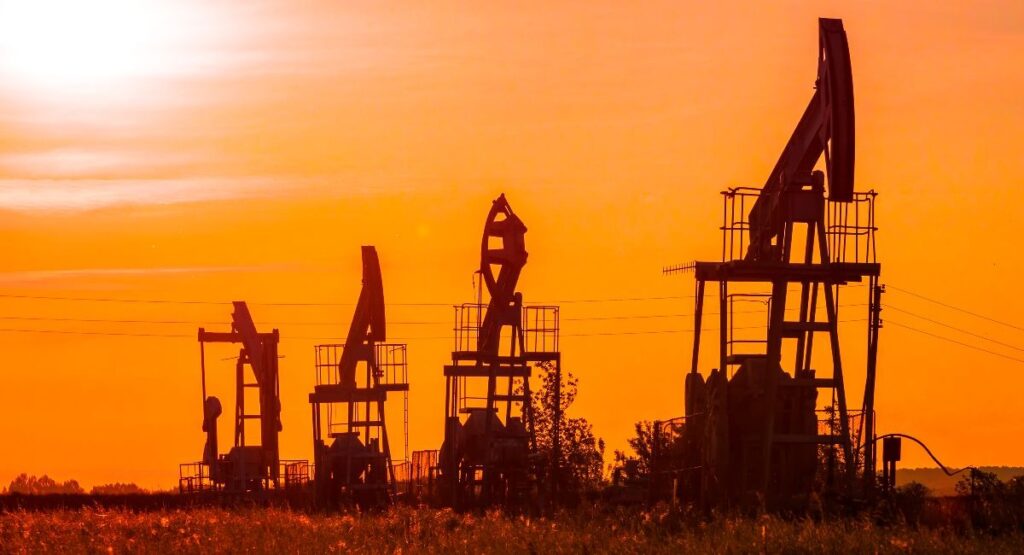 A scenic view of oil pumps silhouetted against a vibrant sunset sky. The sky is ablaze with shades of orange and yellow, with the dark silhouettes of the mechanical structures standing in contrast.