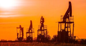 A scenic view of oil pumps silhouetted against a vibrant sunset sky. The sky is ablaze with shades of orange and yellow, with the dark silhouettes of the mechanical structures standing in contrast.